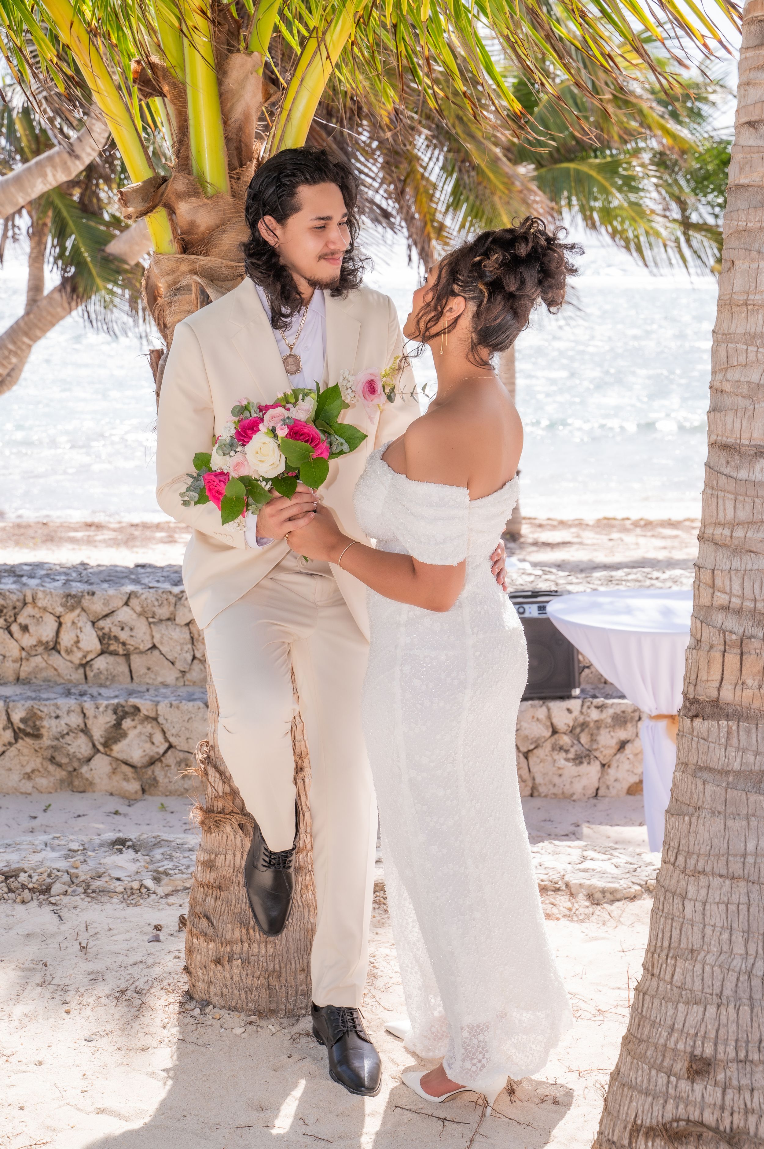 Bride and groom share a romantic moment under palm trees on a tropical beach, bride holding pink and white bouquet.