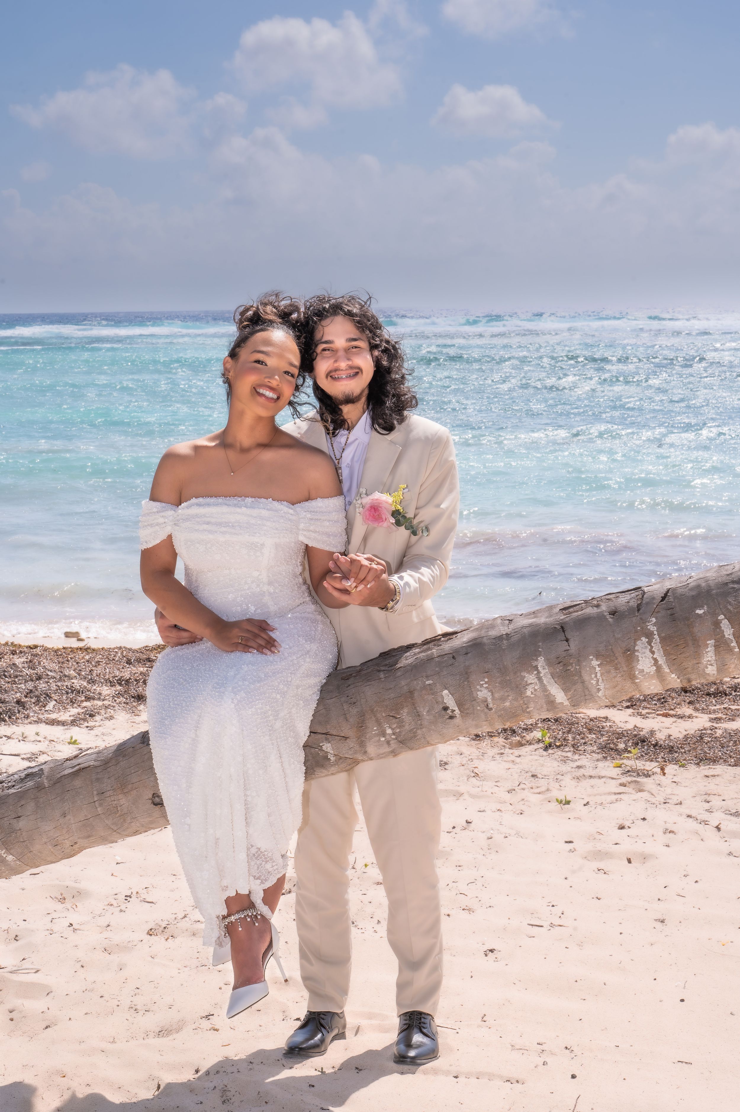 Happy couple in wedding attire posing on a palm tree trunk on a tropical beach with turquoise ocean waters.