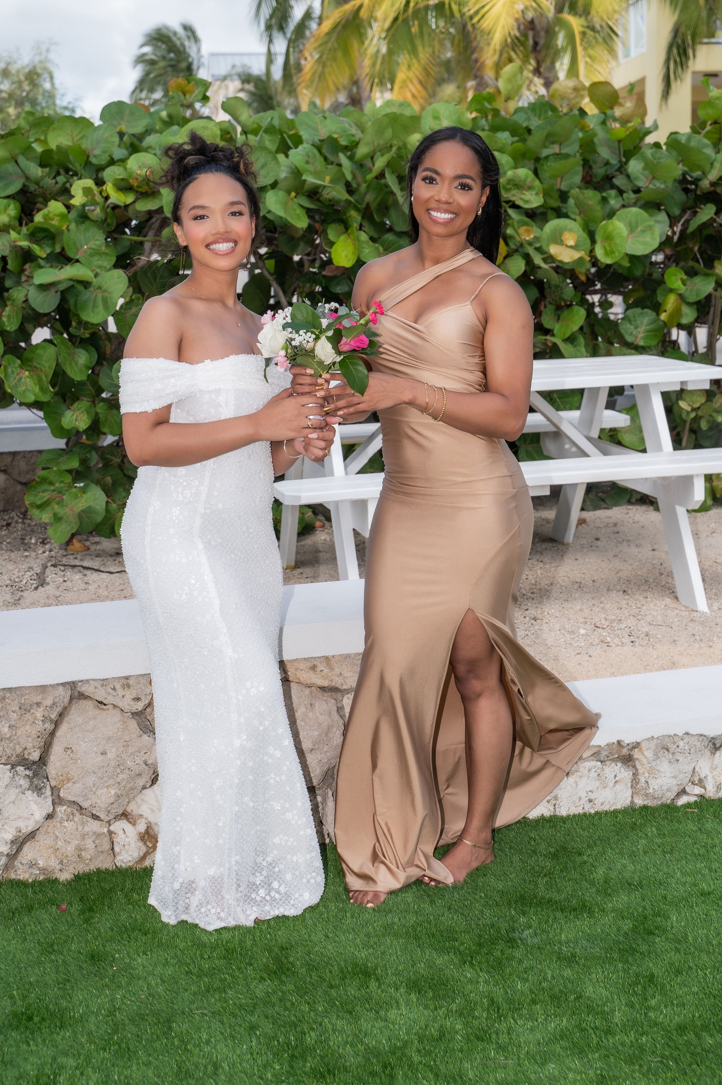 Two women in elegant gowns, one in white lace and one in gold satin, pose with bouquets at an outdoor wedding.