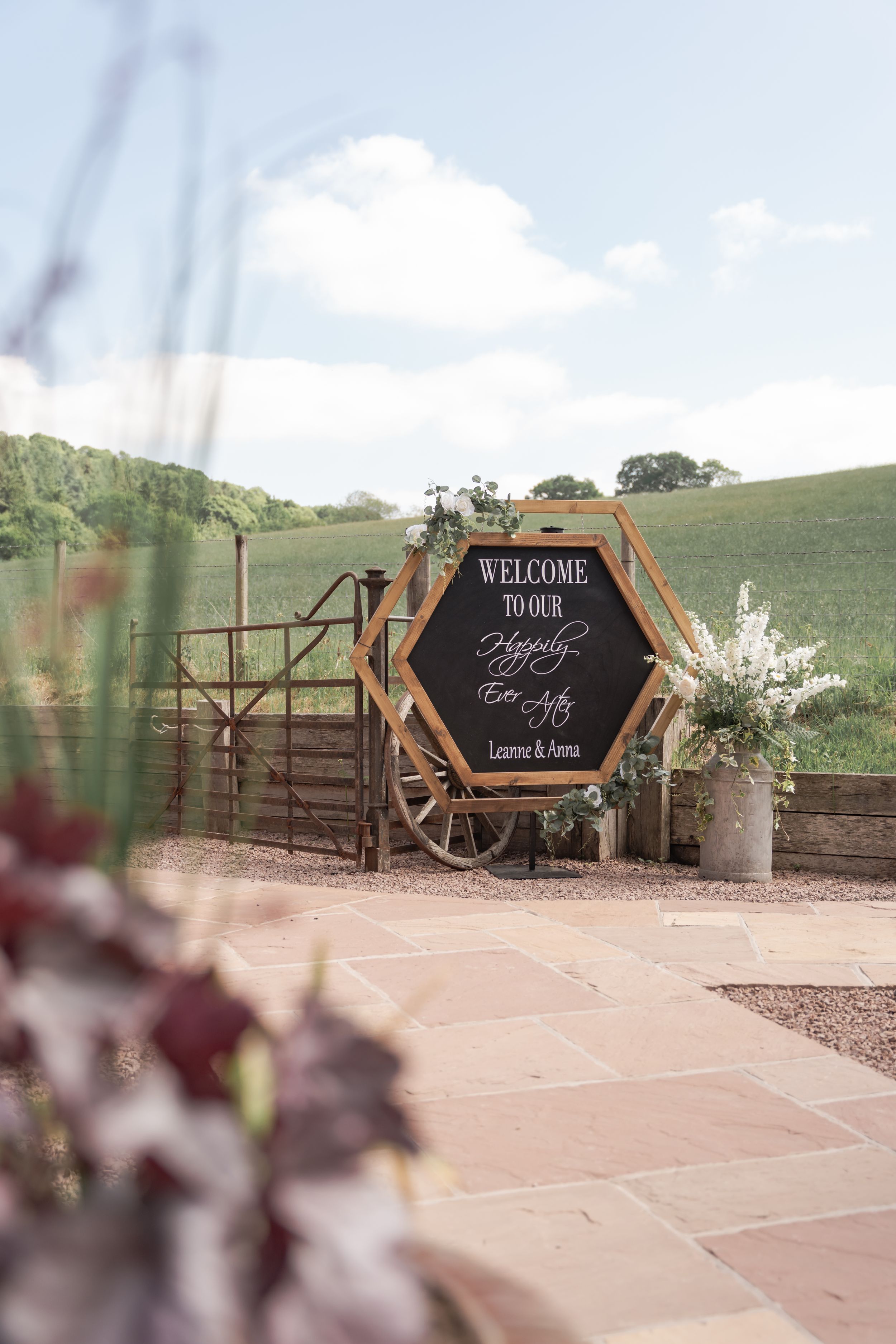 Hexagonal welcome sign with rustic wooden frame and floral decorations sits beside a gate on a countryside path.