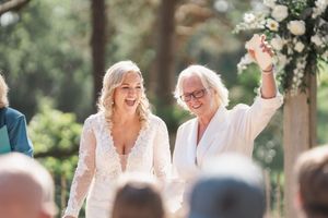 Two people share a joyful moment during an outdoor wedding ceremony under a floral arch.