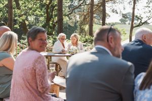 Wedding guests seated at outdoor tables during a forest reception with natural lighting filtering through pine trees.