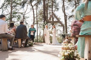 Wedding guests sit outdoors under tall trees during a seaside ceremony on a sunny day.