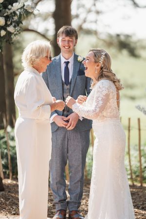 Three people sharing a joyful moment outdoors during a wedding ceremony in white and gray formal attire.