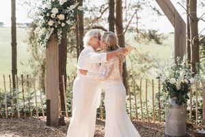 A couple shares a tender moment under a wooden pergola decorated with white floral arrangements and greenery.