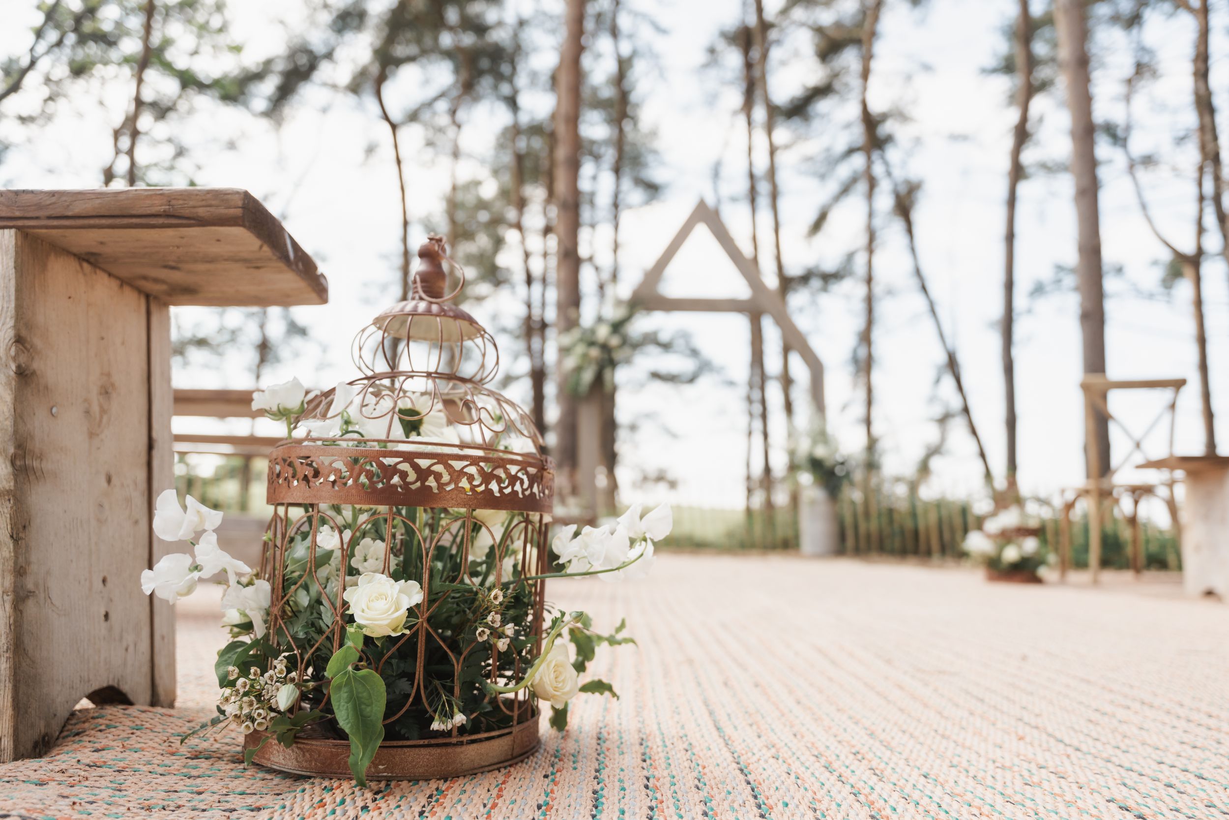 A rustic lantern decorated with delicate white flowers sits beneath a wooden arch in a pine forest wedding setting.