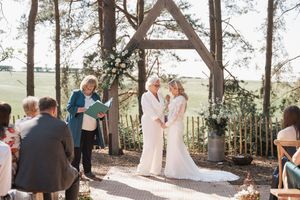 Two people exchanging vows during an outdoor ceremony under a wooden arch surrounded by pine trees and guests.