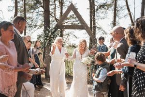 Wedding celebration with couple and guests gathered under rustic wooden arch structure in sunlit forest setting.