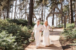 Two people in white dresses walking together on a scenic forest path near the ocean.