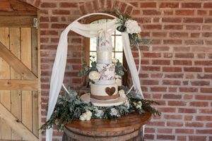 Elegant wedding cake with white floral details and heart accents displayed on wooden table against rustic brick wall.