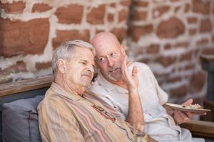 Two elderly friends share a tender moment while sitting together on a couch against a rustic brick wall.