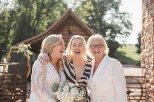 Three women in formal attire laughing together outdoors with rustic building in background.
