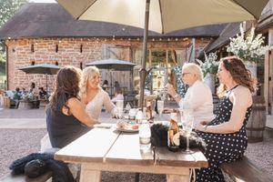 Groups of friends enjoying outdoor dining under an umbrella at a rustic wooden picnic table on a patio.