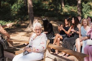Guests seated on wooden benches at an outdoor wedding ceremony while taking photos in a wooded setting.