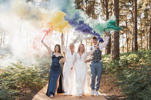 People celebrate in a forest setting with colorful smoke bombs creating a vibrant rainbow effect along a woodland path.