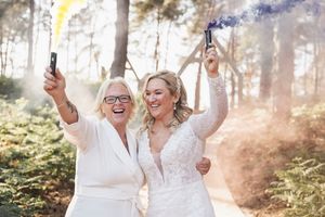 Two brides celebrate joyfully together in a sunlit forest setting with champagne bottles.