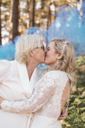 A tender kiss between newlyweds with blue smoke effects in a forest backdrop.