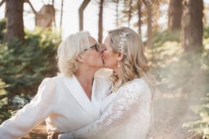 A romantic wedding moment between two brides sharing a kiss in a sunlit forest setting.