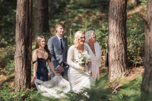 A wedding party gathers in a peaceful woodland setting among tall pine trees on a sunny afternoon.
