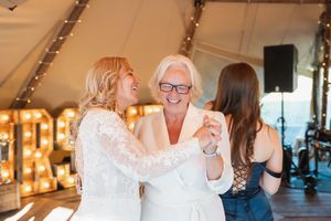 Three people share a joyful moment while dancing together at an indoor reception with string lights in the background.