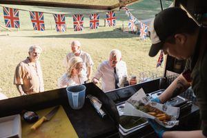 People gather at an outdoor event with British Union Jack bunting while a vendor serves food from a booth.