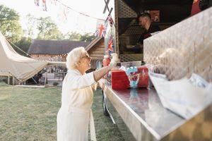 Elderly person decorating at an outdoor celebration with festive bunting and tents in the background.