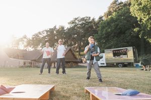 People playing cornhole on a grassy lawn at sunset with a camper van in the background.