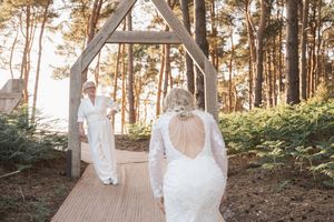 A couple walks down a wooden boardwalk path surrounded by tall pine trees during a golden sunset ceremony.