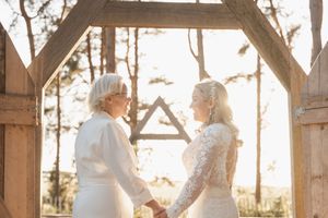 Two people in white lace wedding dresses hold hands during an outdoor ceremony at sunset under a wooden arch.