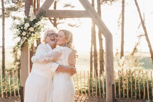 Two brides embrace under a rustic wooden wedding arch adorned with white floral arrangements at sunset.