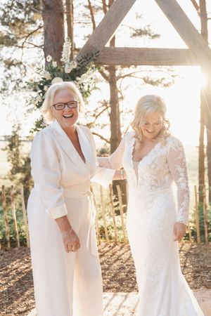 Two people in elegant white outfits share a joyful moment at an outdoor wedding ceremony under a wooden arch at sunset.