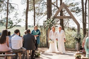 An outdoor wedding ceremony takes place under a wooden triangular arch in a sunlit forest setting.