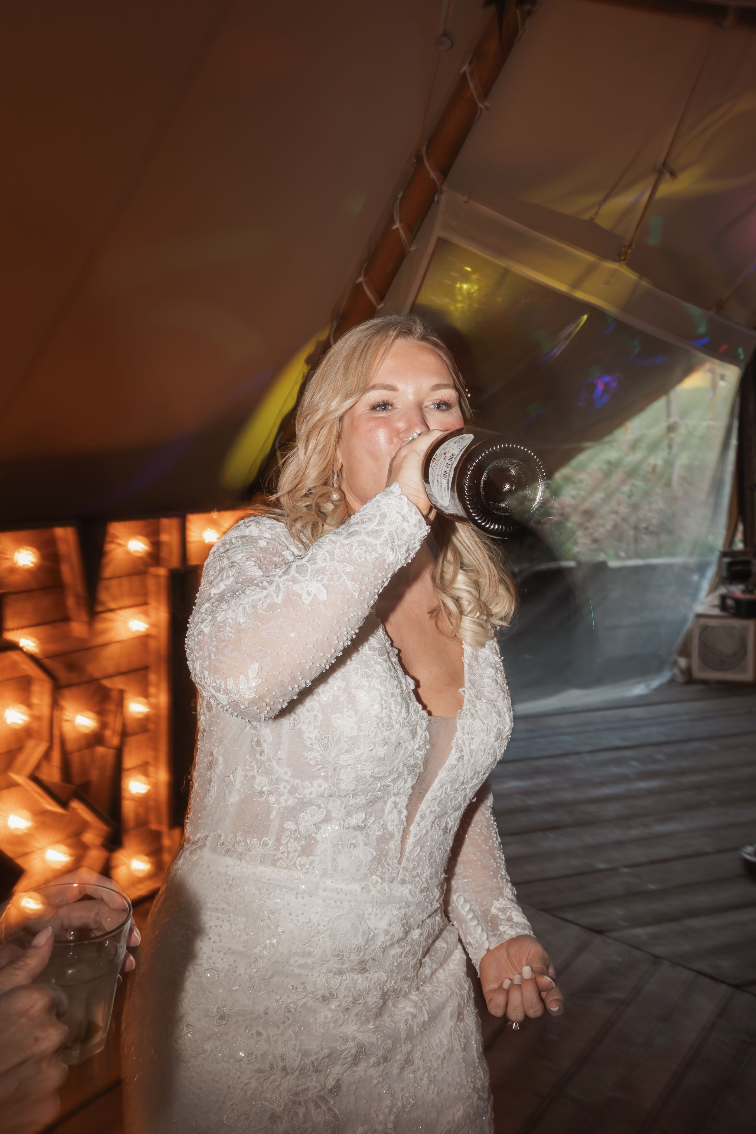 A person in a white lace wedding dress drinks from a water bottle during a celebration with illuminated decor.