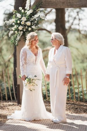 Two figures wearing elegant white dresses share a joyful moment beneath a floral arch during an outdoor celebration.