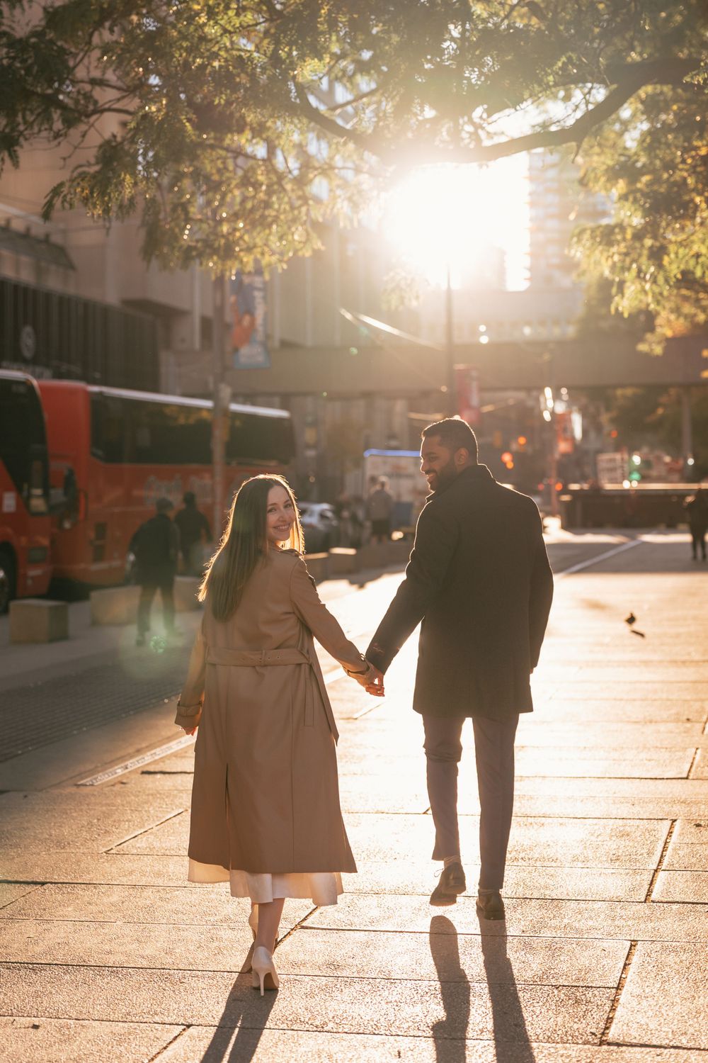 Couple holding hands walking on sunny city street, woman in beige coat, man in black coat, golden backlight.