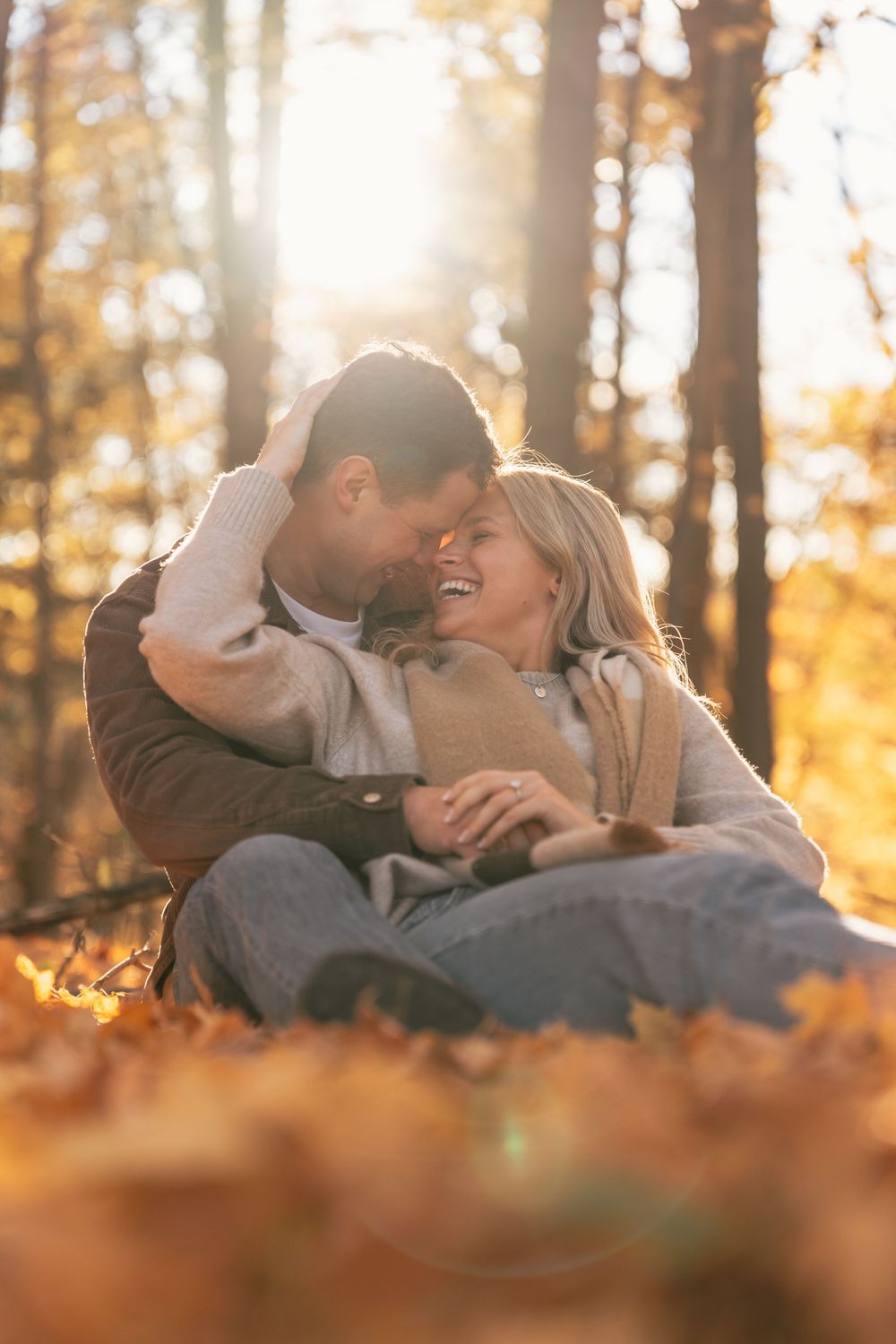 Couple sitting in autumn leaves in a sunlit forest, smiling and embracing romantically during fall season.