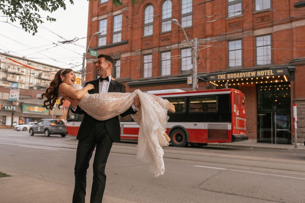 Groom carries bride in wedding attire on urban street with red bus and brick hotel building in background.