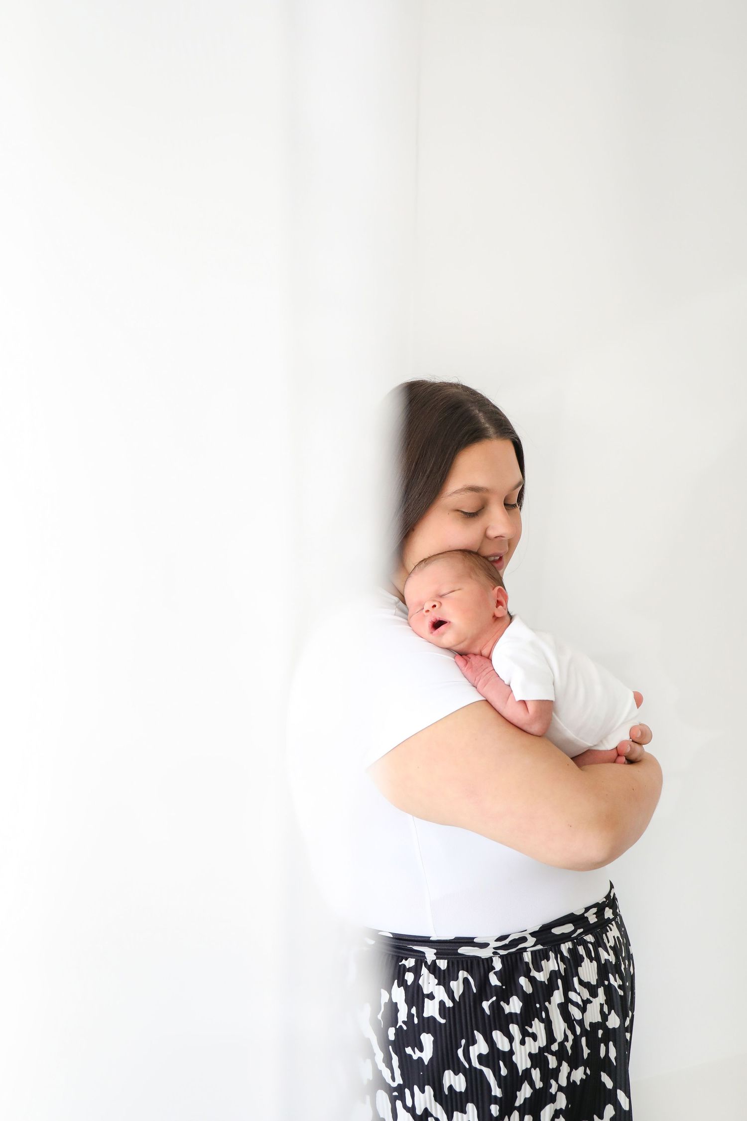 Parent in white top and floral skirt holds sleeping newborn against white wall in minimalist portrait, during newborn shoot in Nottingham