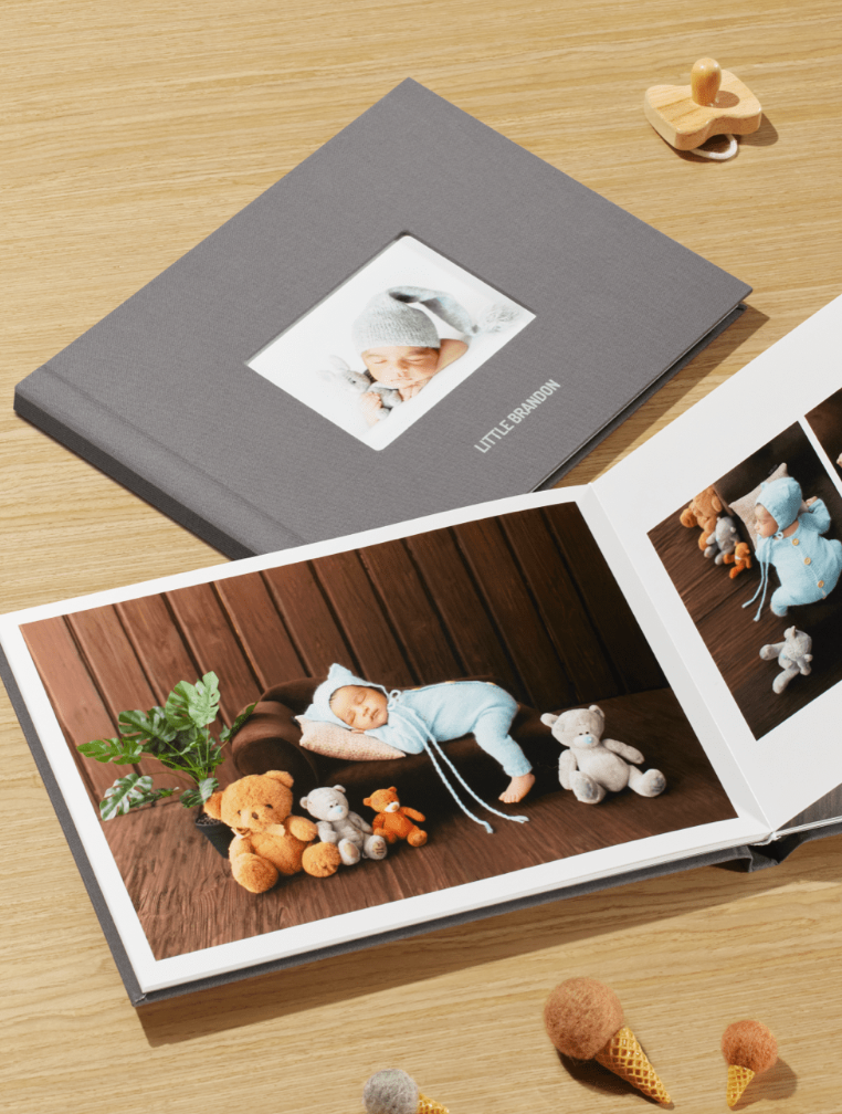 Gray hardcover baby photo book open showing infant in blue outfit with stuffed animals, beside closed album on wood surface. Phone Book