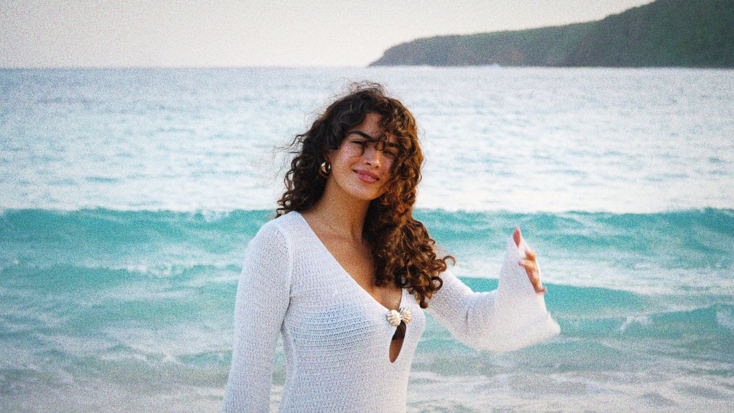 Person in white sweater standing on beach with turquoise ocean waves and coastal hills in background.