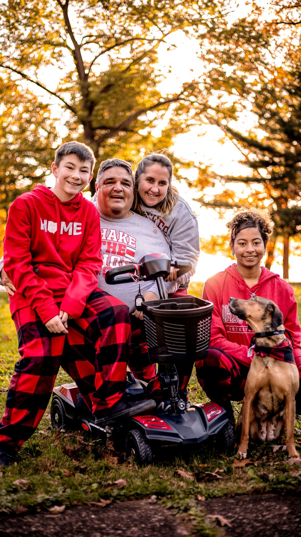 Family wearing red and buffalo plaid pajamas poses together outdoors with their dog during golden autumn sunset.