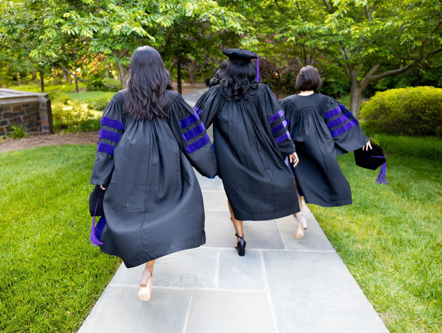 Graduates wearing black caps and gowns walk together on a garden path during commencement ceremony.