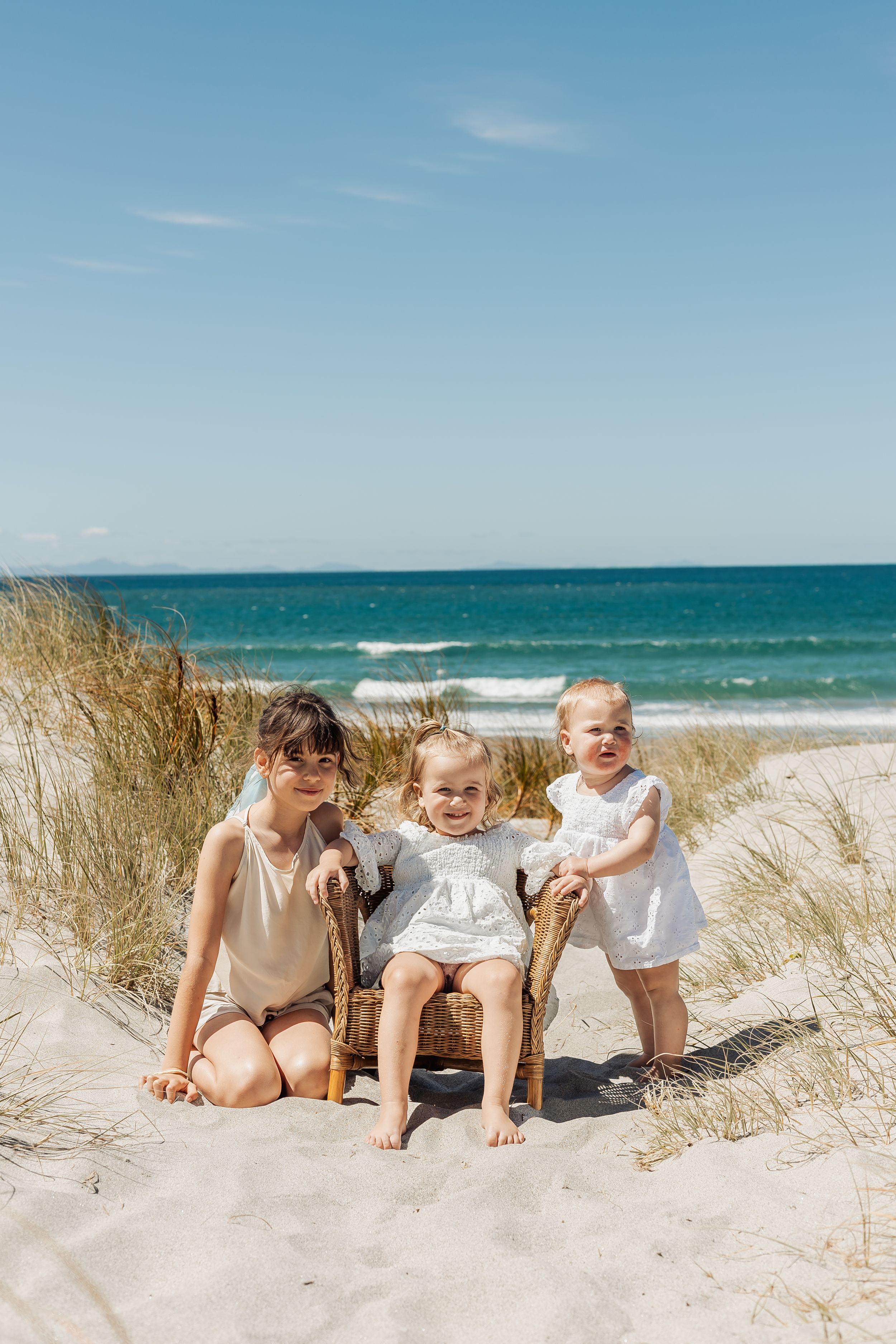 Three young children sitting on sandy beach dunes with turquoise ocean waves and blue sky backdrop.
