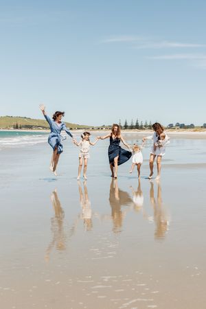 Two women and three children joyfully playing and jumping on a sunny beach with wet sand reflections.