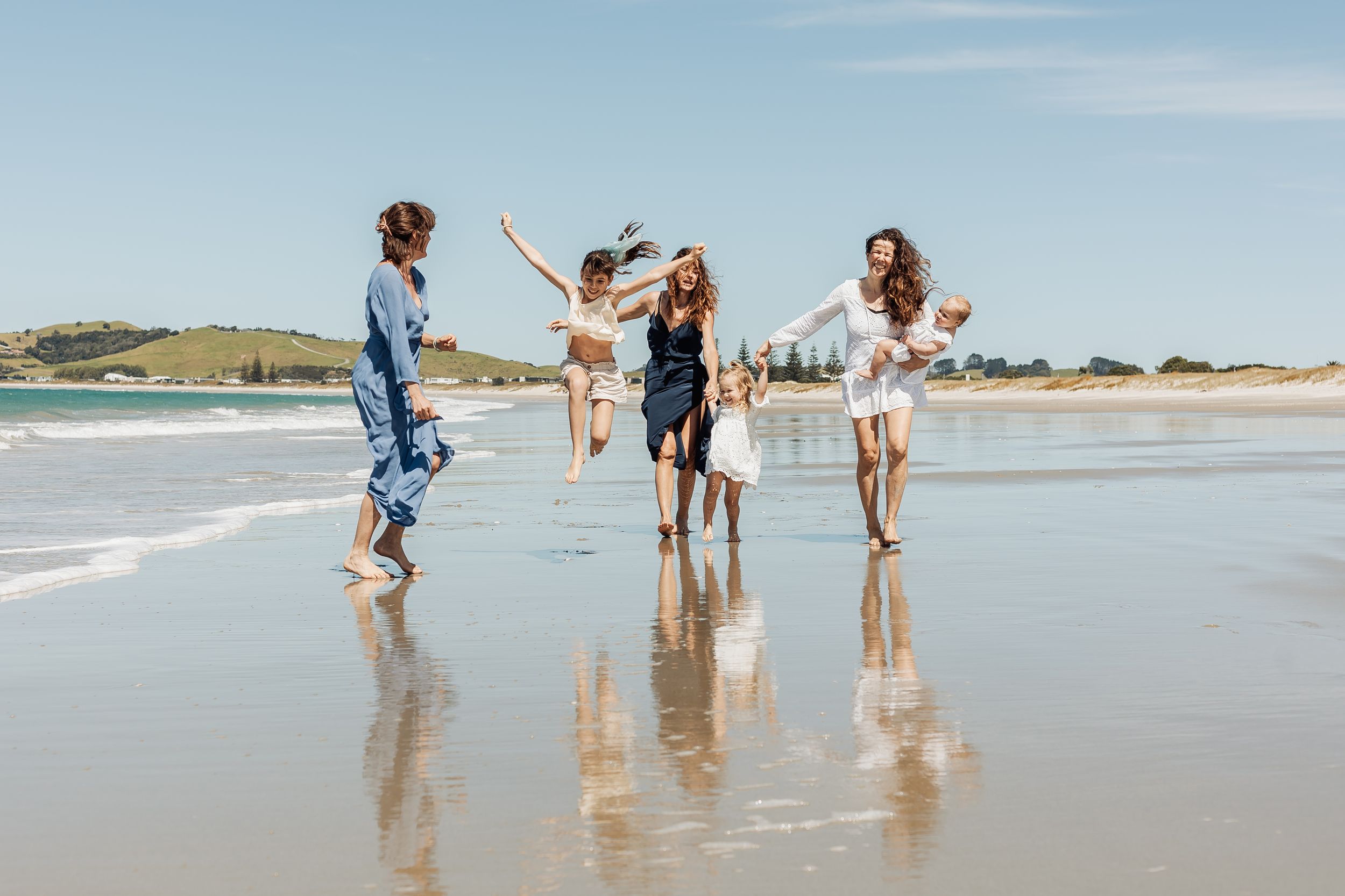 Group of friends jumping and laughing on a sunny beach with calm waves and green hills in the background.