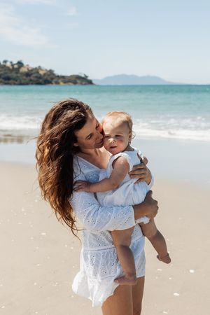 A mother in a white dress holds her baby on a sunny beach with turquoise ocean waves in the background.