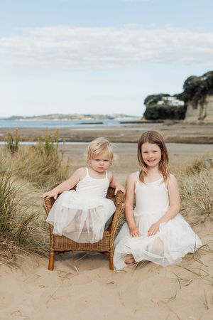 Two young girls in white dresses pose on sandy beach, one sitting in wicker chair, smiling at camera.