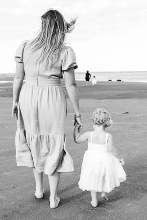 Black and white photo of mother and young daughter holding hands walking on beach, both in dresses, viewed from behind.