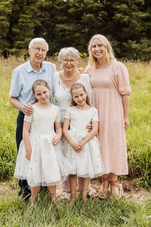 Multi-generational family portrait with grandparents, mother, and two girls in white dresses outdoors.