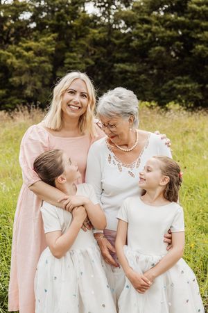 Three generations of women, grandmother, mother, and two girls in white dresses, smiling outdoors in a green meadow.